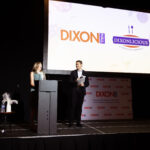 A woman speaks at a podium while a man stands beside her holding papers on a stage with a large screen displaying "Dixon Hall" and "Dixonlicious."
