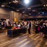 A large crowd of people gathered in an event space, some standing and some seated on couches, attentively watching a presentation.
