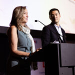 A woman speaks at a podium while a man stands beside her, attentively listening.