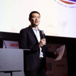 A man in a dark blazer speaks into a microphone while holding papers on stage.