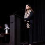 A woman in a black outfit with a patterned scarf speaks at a podium on stage.