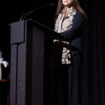 A woman in a black outfit with a patterned scarf speaks at a podium on stage.