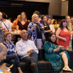 A group of people, some seated and some standing, attentively watching an event while enjoying food and drinks.