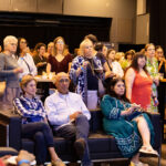 A diverse audience, both seated and standing, attentively watching an event while holding food and drinks.