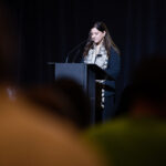 A woman stands at a podium, delivering a speech on a dimly lit stage, with audience members visible in the foreground.