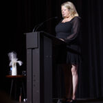 A woman in a black dress stands at a podium, delivering a speech on a dimly lit stage.