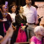 A group of people attentively watching an event, with a woman in a black jacket holding a pink bag in the foreground.