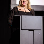A woman in a black dress speaking at a podium on stage.