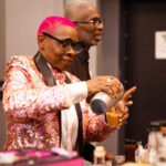 A bartender with bright pink hair and a shiny patterned jacket pours a drink from a shaker, with another person standing behind them.