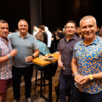Four men stand together at a high-top table, smiling and enjoying drinks and food at a social event.