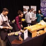 Two men wearing aprons prepare and serve small food samples at a tasting station, with promotional banners and other attendees in the background.