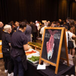 A food station with chefs preparing dishes, featuring a chalkboard sign with a drawing of a fish labeled "KING," and a crowd of attendees in the background.