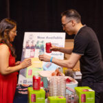 A vendor in a black shirt handing a canned beverage to a woman in a red dress at a tasting station, with promotional signage and product displays in the background.