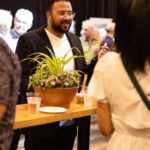 A man in a black blazer and white shirt is engaged in conversation with a woman at a high-top table with drinks and a potted plant, surrounded by other event attendees.
