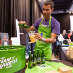 A man wearing a green Steam Whistle apron and a purple shirt pours beer from a bottle into a plastic cup at a beverage station, with branded merchandise and bottles displayed on the table.