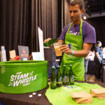 A man wearing a green Steam Whistle apron and a purple shirt pours beer into a plastic cup at a branded beverage station, with bottles and merchandise displayed on the table.