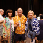 Three people pose together at an event, with the man in the center wearing a bright orange shirt and holding a can of Olé beverage, while the two women on either side wear colorful patterned outfits.