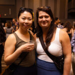Two women pose together at an event, both smiling at the camera. One holds a drink while the other carries a crossbody bag. The background shows other attendees mingling.