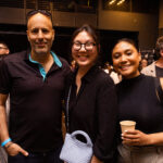 Three people pose together at an event, smiling at the camera. One person holds a drink, while another carries a woven handbag. The background shows other attendees mingling.