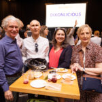 Four people pose together at an event, standing around a table with food and drinks. A large screen in the background displays "DIXONLICIOUS."