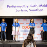 Four children in blue "Dixon Hall Music School" shirts perform on stage with microphones. A large screen above displays their names.