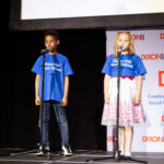 Two children in blue "Dixon Hall Music School" shirts stand on stage with microphones, performing in front of an audience.
