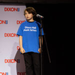 A child in a blue "Dixon Hall Music School" shirt stands on stage in front of a microphone, ready to perform.
