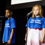 Two young children in blue "Dixon Hall Music School" shirts stand on stage, each in front of a microphone, performing.