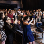 A large audience gathers at an indoor event, with many people capturing the moment on their phones.