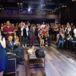 A large audience in an indoor venue claps and takes photos, showing enthusiasm and engagement.