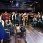 A lively audience in an indoor venue claps and takes photos, showing engagement and excitement.