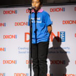 A young performer in a blue Dixon Hall Music School shirt sings into a microphone on stage.