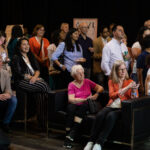 A diverse group of people, some seated and others standing, attentively watching an event while some take photos.