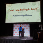 A young performer sings on stage under a large screen displaying "Can't Help Falling in Love" by Elvis Presley, performed by Marcus.