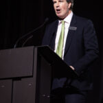 A man in a suit and tie speaks at a podium on a dimly lit stage.