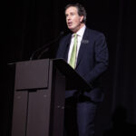 A man in a dark suit and green tie speaks at a podium on a dimly lit stage.