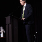 A man in a dark suit and green tie speaks at a podium on a dimly lit stage. A decorative vase with white feathers sits on a small table nearby.