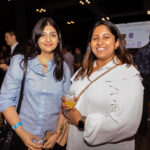 Two women smile at the camera while holding drinks at a social event. The background is filled with other attendees and a presentation screen.
