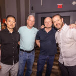 Four men pose together at an indoor social event, smiling at the camera. The background features a door with an exit sign.