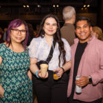 Three people smile at the camera while holding drinks at a social event, surrounded by other attendees in a lively indoor setting.