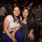 Two women sit together on a black couch, smiling at the camera at a social event.
