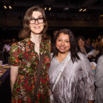 Two women pose together at a social event, smiling at the camera. One wears glasses and a floral dress, while the other wears a silver outfit with a crossbody bag.