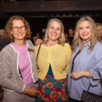 Three women pose together at a social event, smiling at the camera. They are dressed in colorful and casual outfits, with one wearing glasses and a name tag.