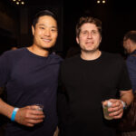 Two men stand side by side at a social event, smiling at the camera while holding drinks. They are dressed casually, one wearing a navy blue shirt and the other in black.