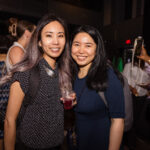 Two women smile at the camera at a social event. One is holding a drink, and both are wearing backpacks. They are dressed in casual attire.