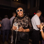 A stylish woman wearing sunglasses and headphones smiles while holding a drink and a phone with a decorative case at a social event.