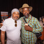 Two men pose together at an event, one wearing a white shirt and headscarf with sunglasses hanging from his shirt, and the other wearing a plaid shirt, beaded necklace, and a hat while holding a drink.