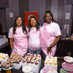 Three women wearing pink aprons stand behind a dessert table filled with cupcakes and macarons, smiling at the camera.