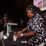 A DJ wearing headphones and a patterned shirt is mixing music on a turntable at an event.