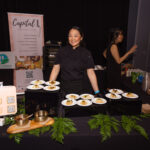 A chef in a black uniform stands behind a table displaying plated dishes at a catering event. A banner for "Capital L" is visible in the background.